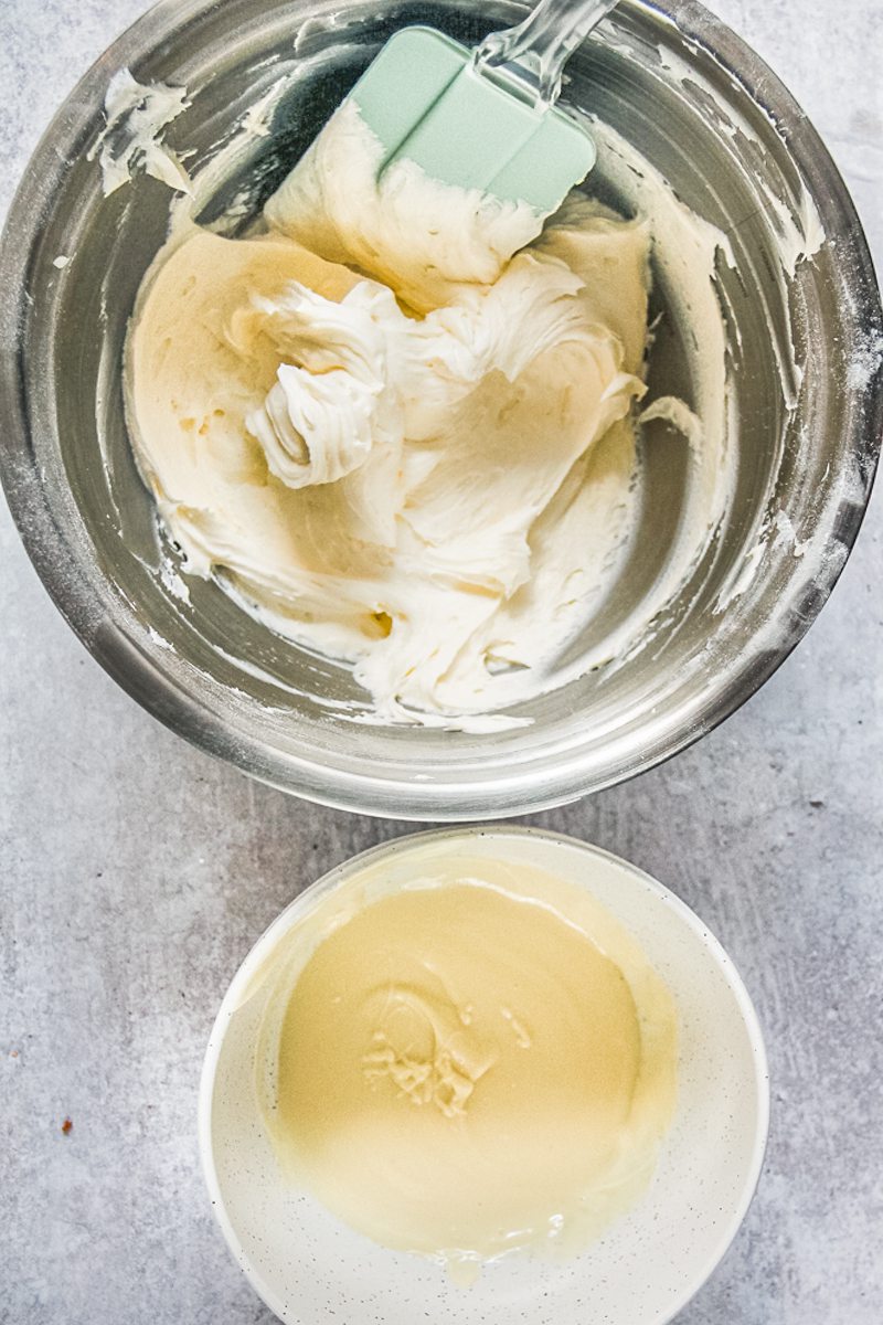 Melted chocolate sits in a white ceramic bowl beside whipped buttercream in a stainless steel bowl on a gray surface.