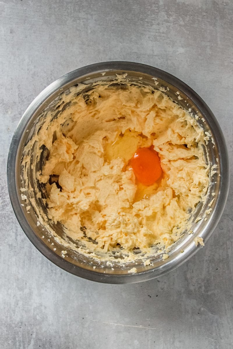 An egg sits on top of whipped butter and sugar in a stainless steel bowl on a gray surface.