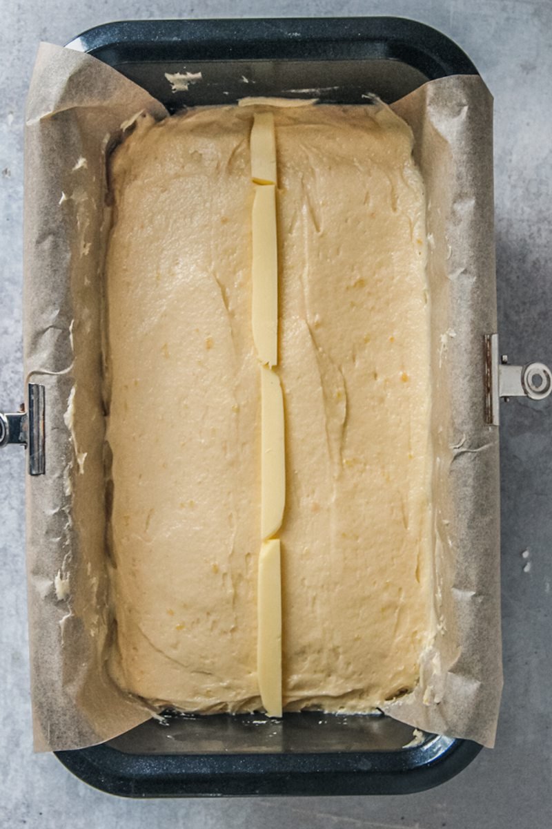 Lemon Drizzle loaf cake sits in a metal loaf tin with thin pieces of butter down the center of the cake on a gray surface.