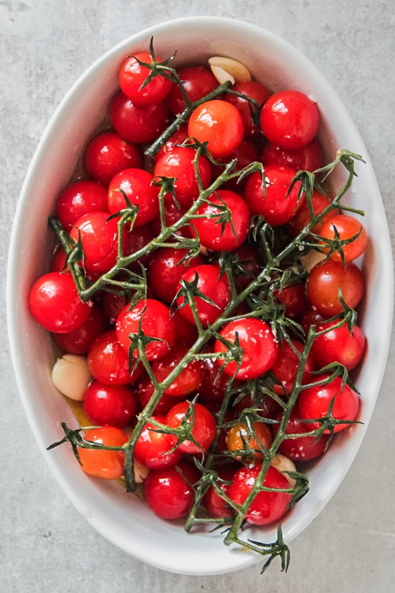 Vine cherry tomatoes with garlic cloves sit in an oval white baking dish on a gray surface.