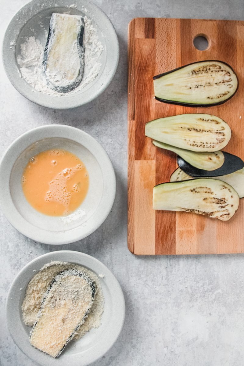 Slices of eggplant sit on a wooden board with other pieces being dredged in bowls of flour, egg and breadcrumbs on a gray surface.