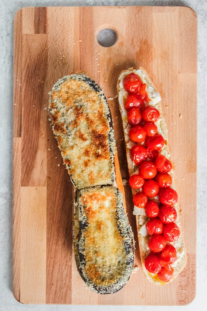 Roasted cherry tomatoes sit on a halved baguette and lightly fried eggplant pieces lay on the other side of a baguette on a wooden board on a gray surface.