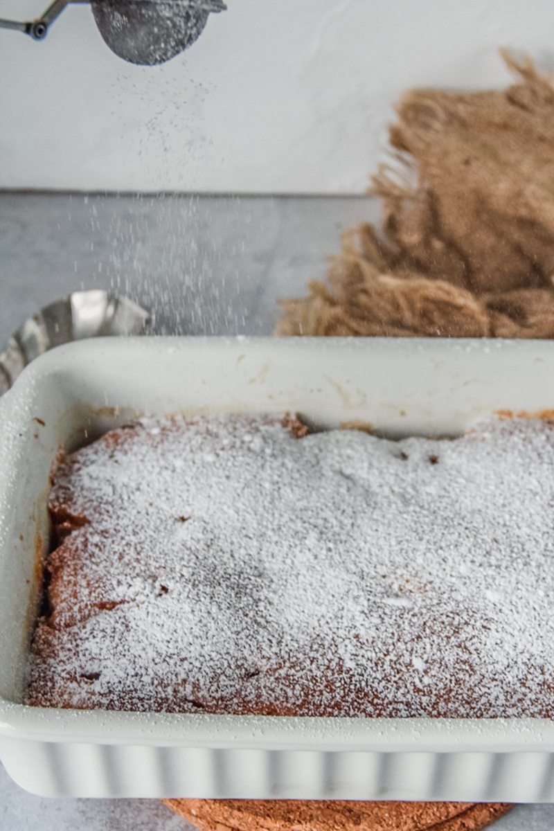 Apple pudding has powdered sugar sprinkled on top as it sits on cork trays on a gray surface.