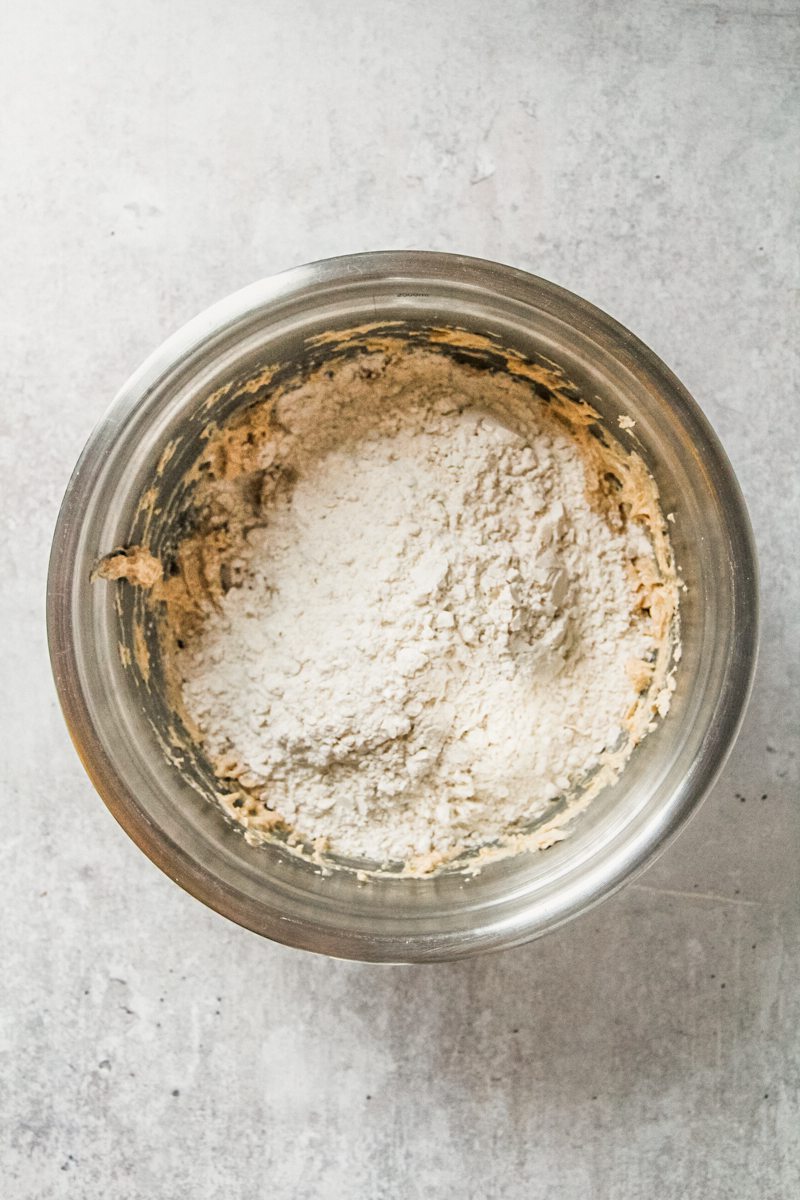 Flour sits on top of a cookie batter in a stainless steel bowl on a gray surface.