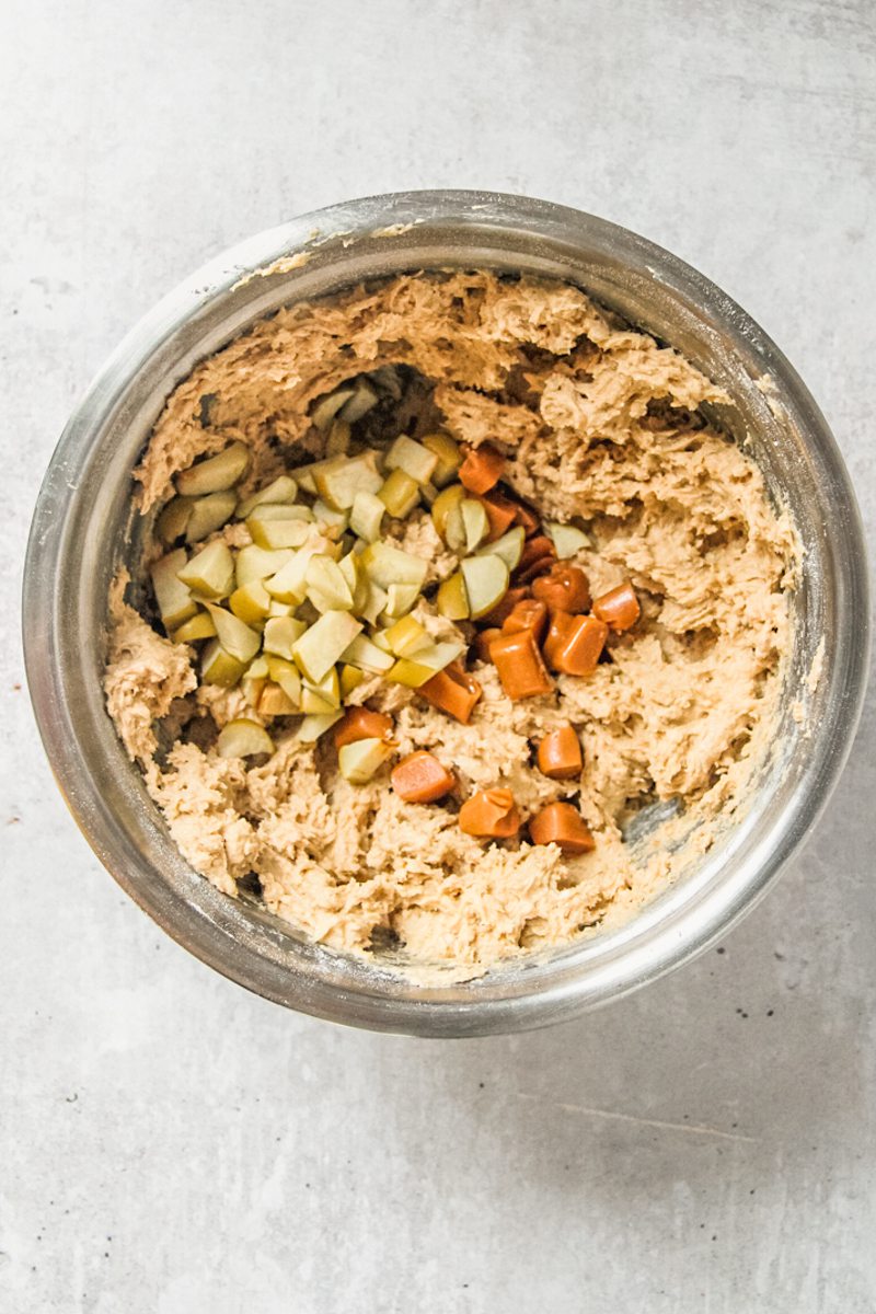 Dried apple pieces and caramel sit on top of cookie batter in a stainless steel bowl on a gray surface.