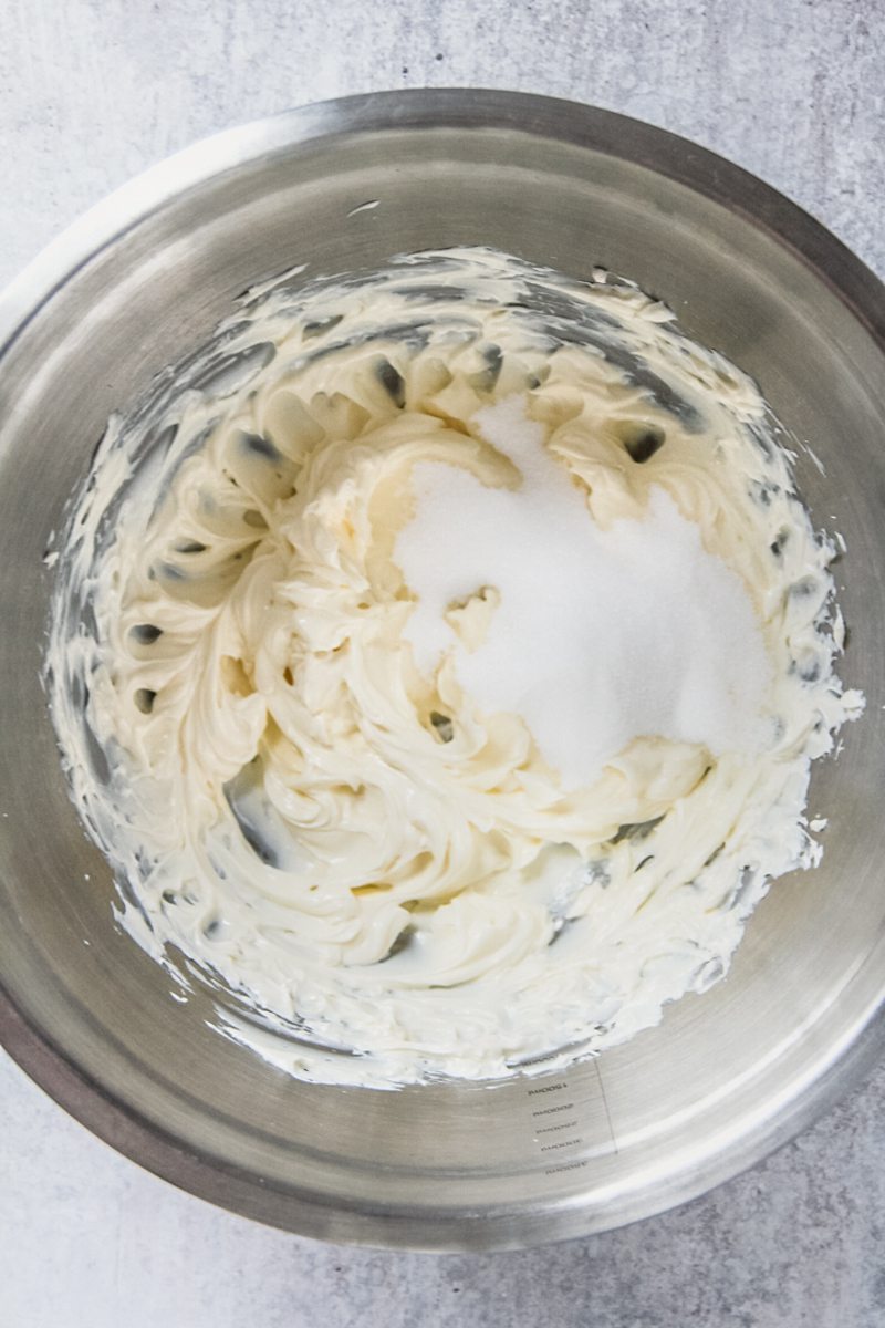 Cream cheese and sugar sit in a stainless steel bowl on a gray surface.