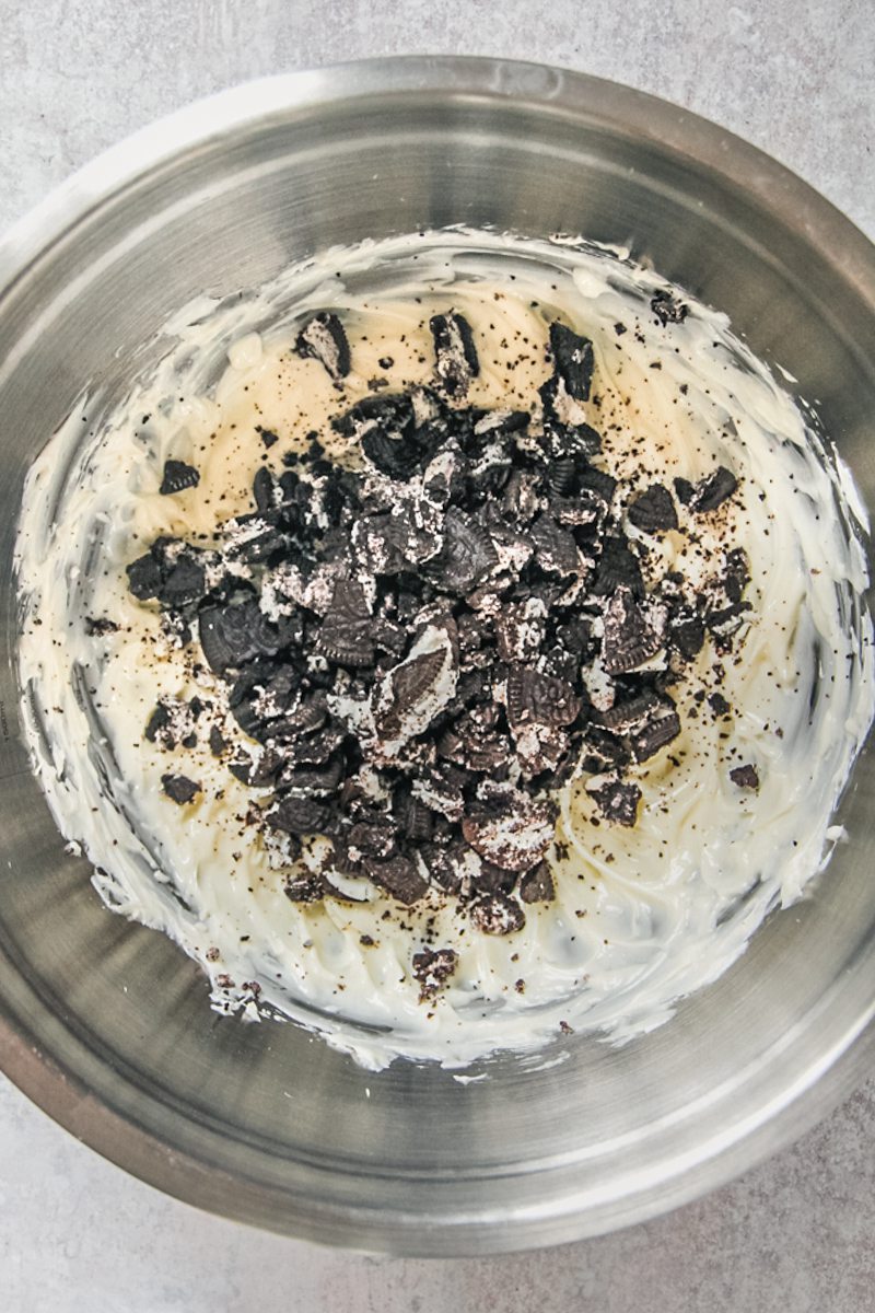 Oreo pieces sit on top of a cheesecake mixture in a stainless steel bowl on a gray surface.