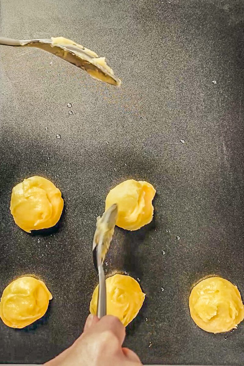 Choux pastry batter is shaped with a teaspoon on a baking tray for a no piping bag recipe.