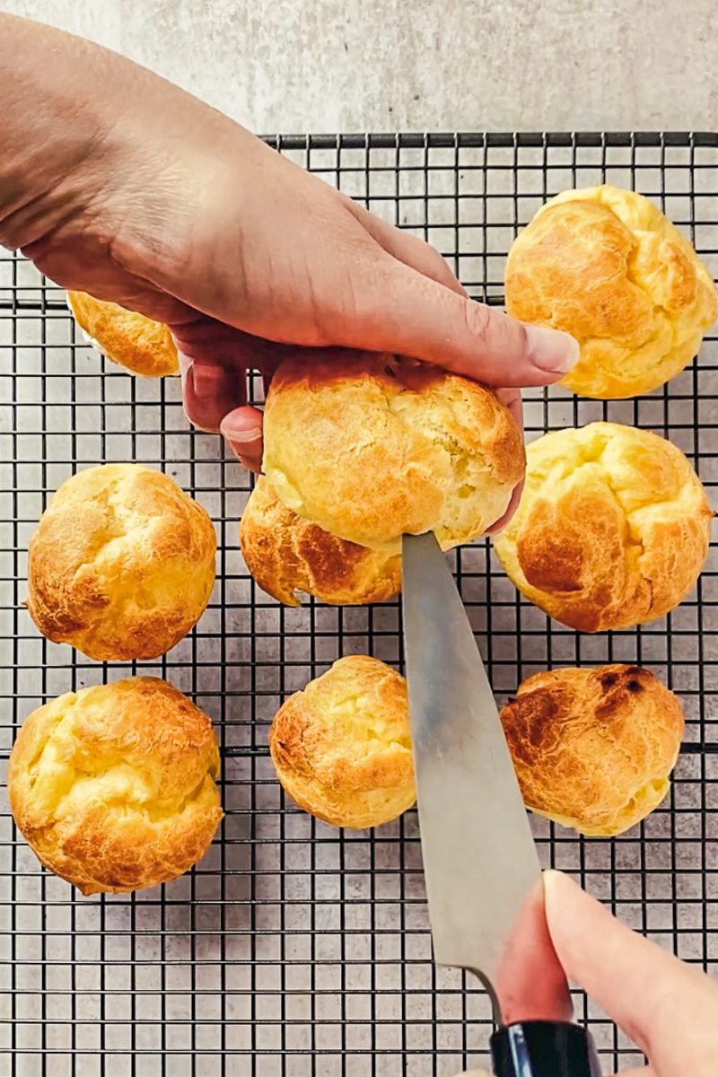 A cream puff is halved with a knife above a cooling rack of cream puffs above a gray surface.