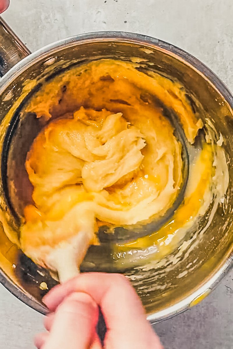 Choux pastry with stirred with a wooden spoon in a stainless steel saucepan on a gray surface.