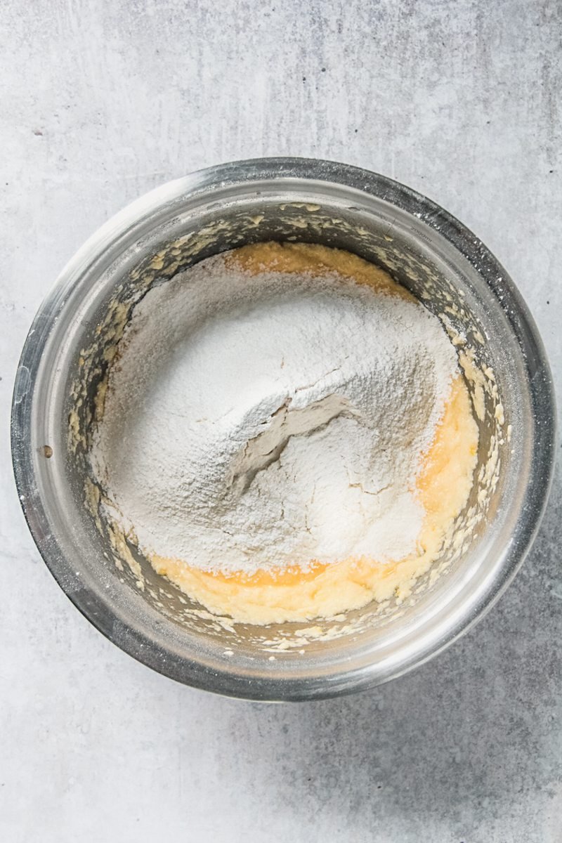 Dry ingredients sit on top of whipped butter and eggs in a stainless steel bowl on a gray surface.