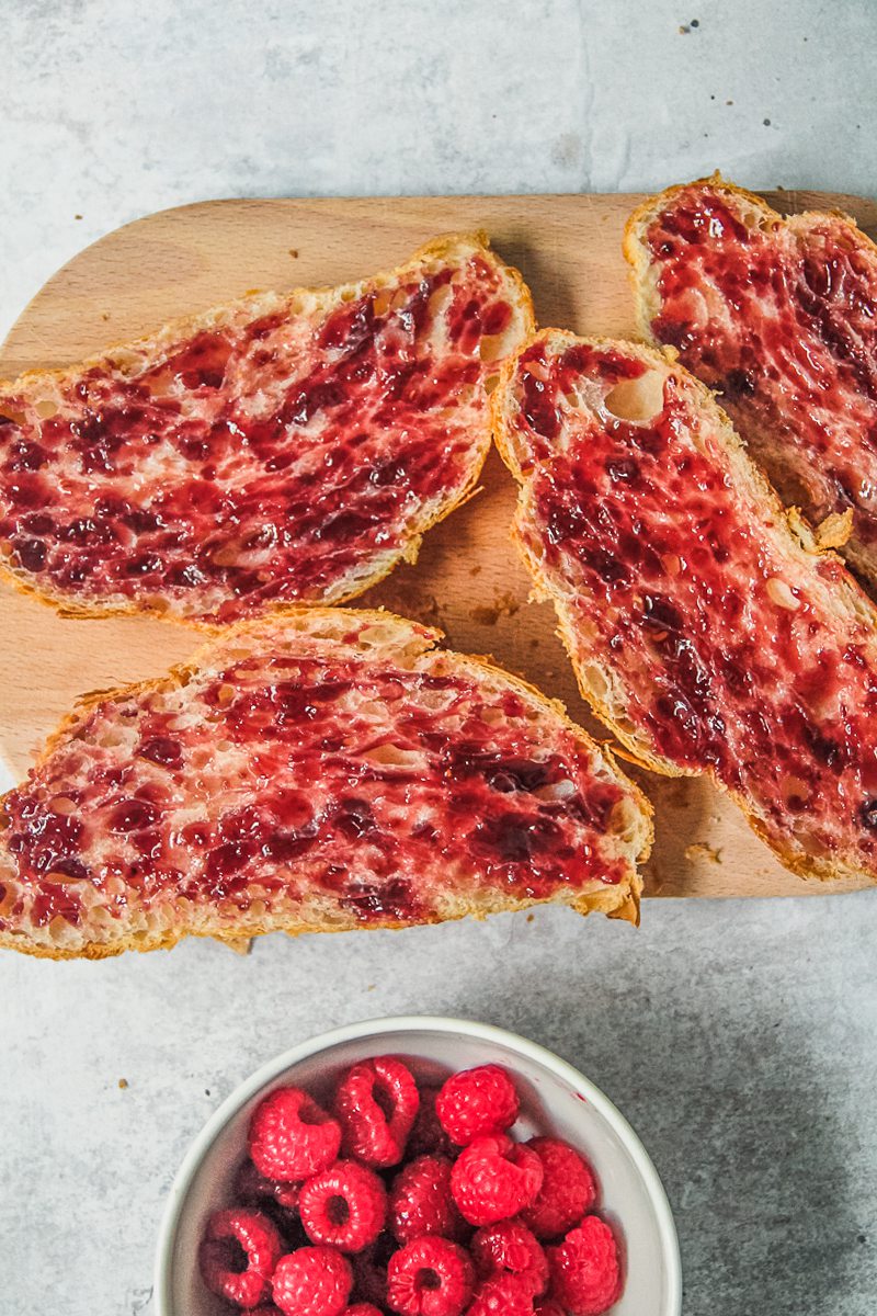 Halved day old croissants topped with raspberry jam sit on a wooden chopping board on a gray surface.