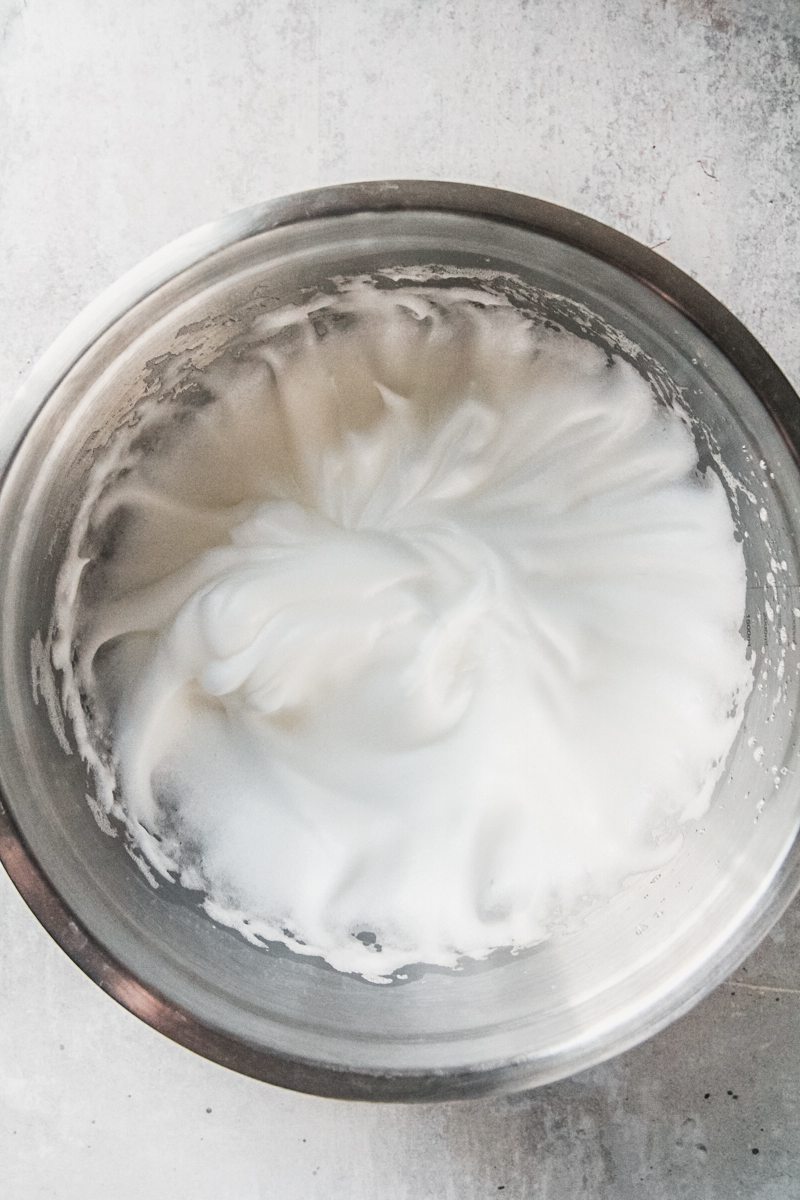 Whipped soft peaks meringue sits in a stainless steel bowl on a gray surface.