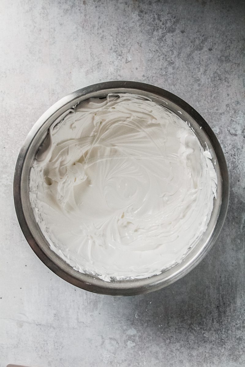 Whipped glossy meringue sits in a stainless steel bowl on a gray surface.