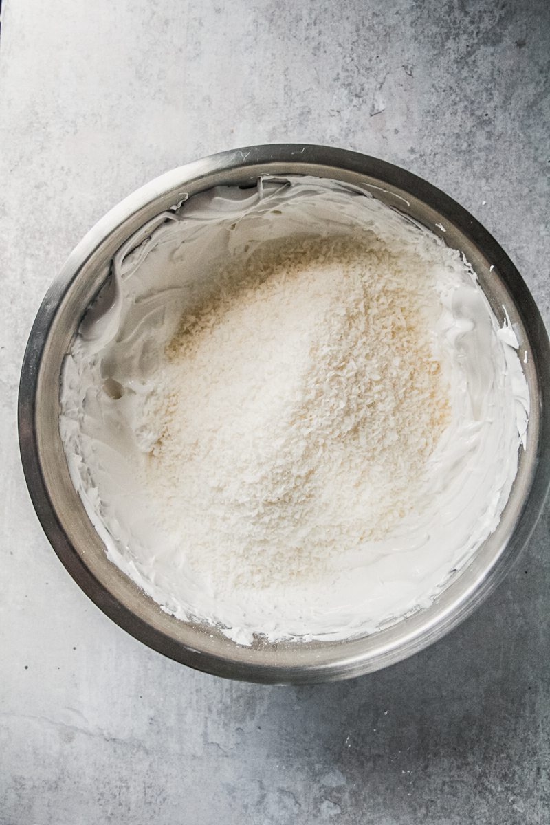 Desiccated coconut sits on top of whipped meringue in a stainless steel bowl on a gray surface.