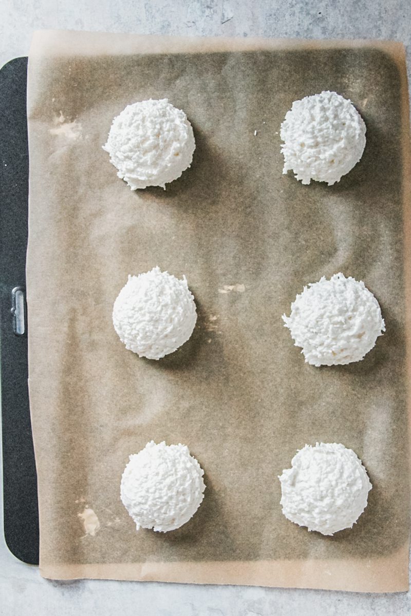 Scoops of unbaked coconut meringue cookies on a parchment paper lined baking tray on a gray surface.