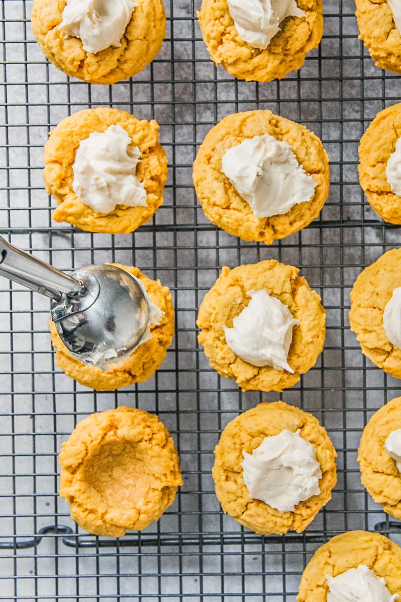 Maple buttercream frosting is placed into the center of pumpkin cookies on a cooling rack above a gray surface.