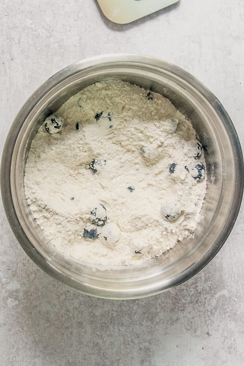 Blueberries and dry ingredients sit stirred together in a stainless steel bowl on a gray surface.