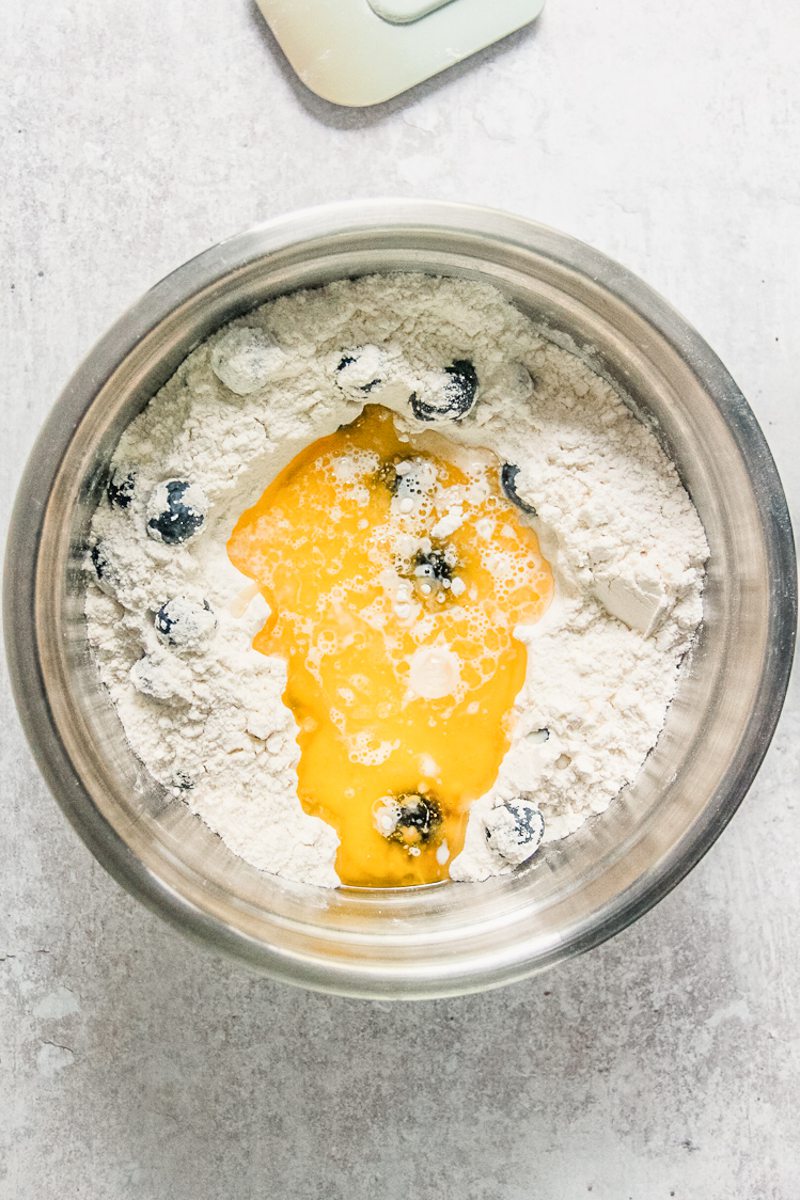 Melted butter sits in the well of a blueberry and dry ingredient mixture in a stainless steel bowl on a gray surface.