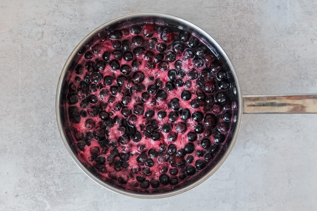 Cooked blueberries sit in a stainless steel pan on a light grey background.