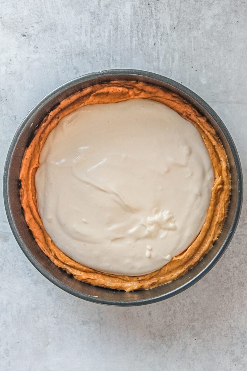 Cheesecake filing sits in the center of the pumpkin cake batter in a round metal tin on a gray surface.