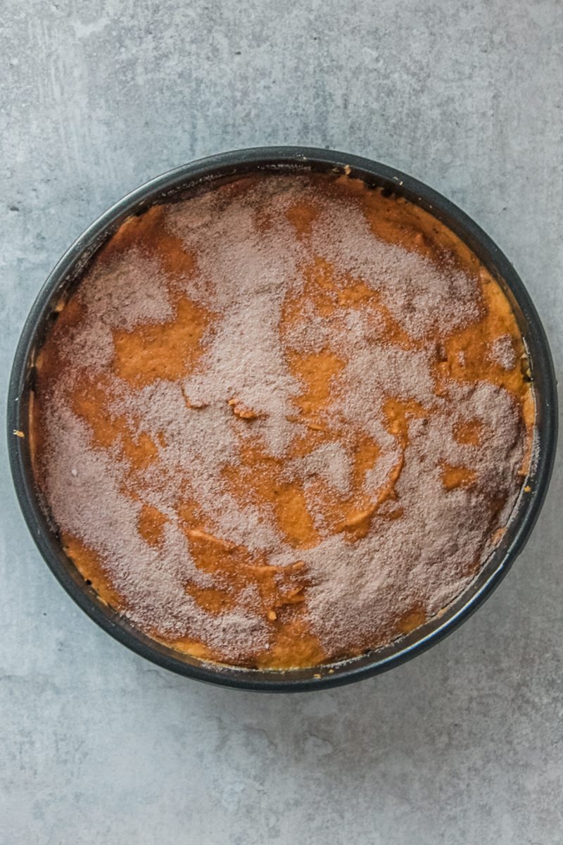 A cinnamon sugar topping sits on top of a pumpkin cake in a round metal tin on a gray surface.