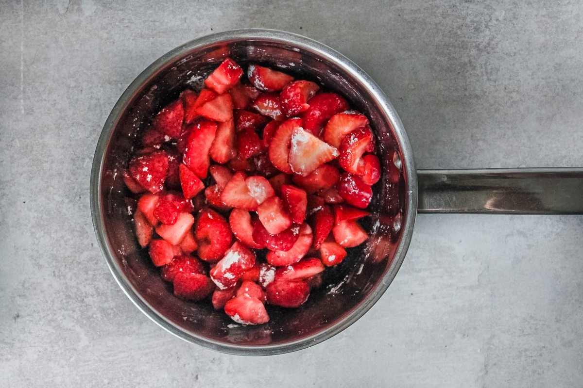 Roughly chopped strawberries sit in a saucepan with sugar and cornstarch on a light grey background.