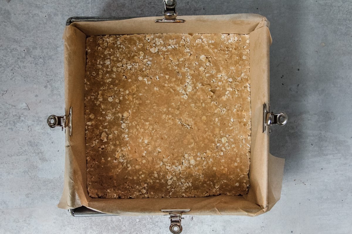 A oat streusel mixture sits pressed evenly into a square baking tin for strawberry bars, sitting on a light grey background.