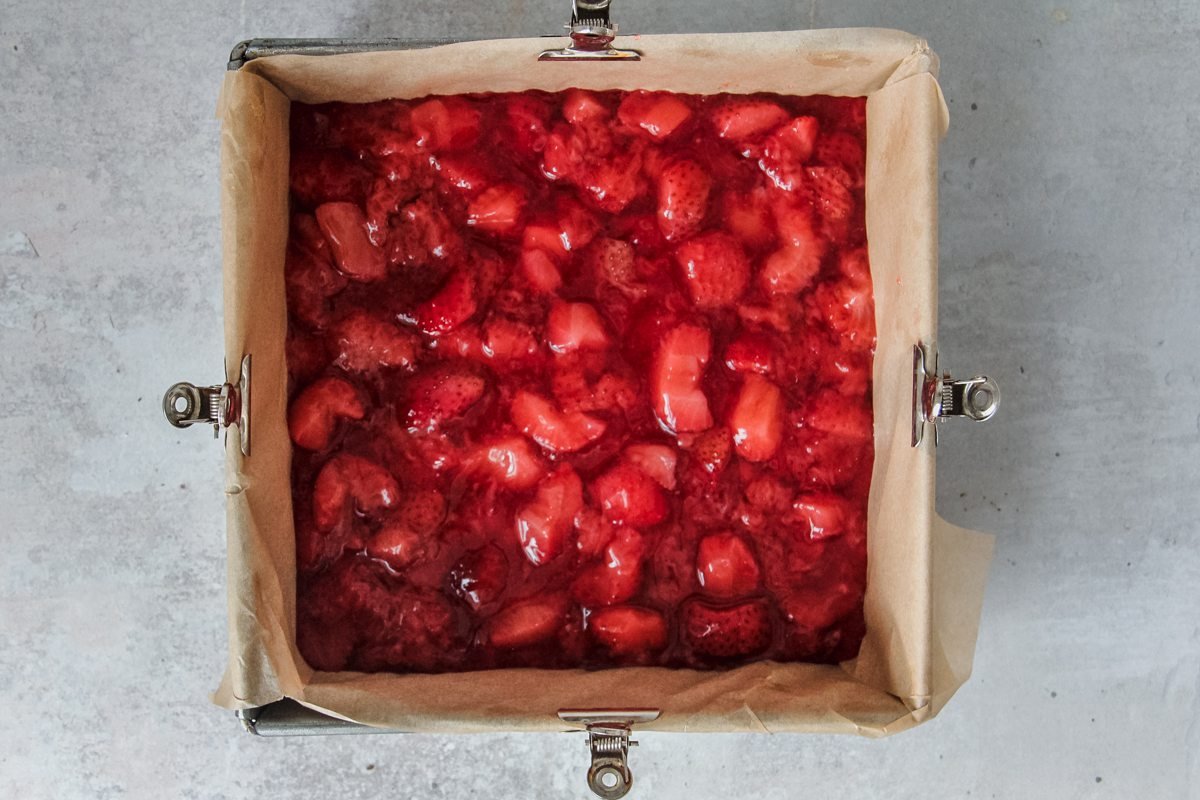 A homemade strawberry jam mixture sits atop a oat streusel base in a square baking tin on a light grey background.