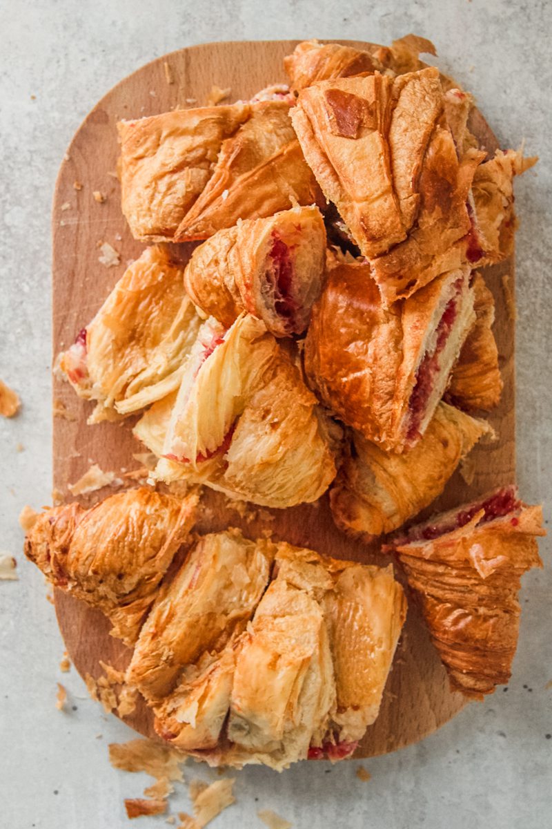 Pieces of day old croissants sit on a rectangle wooden chopping board on a gray surface.