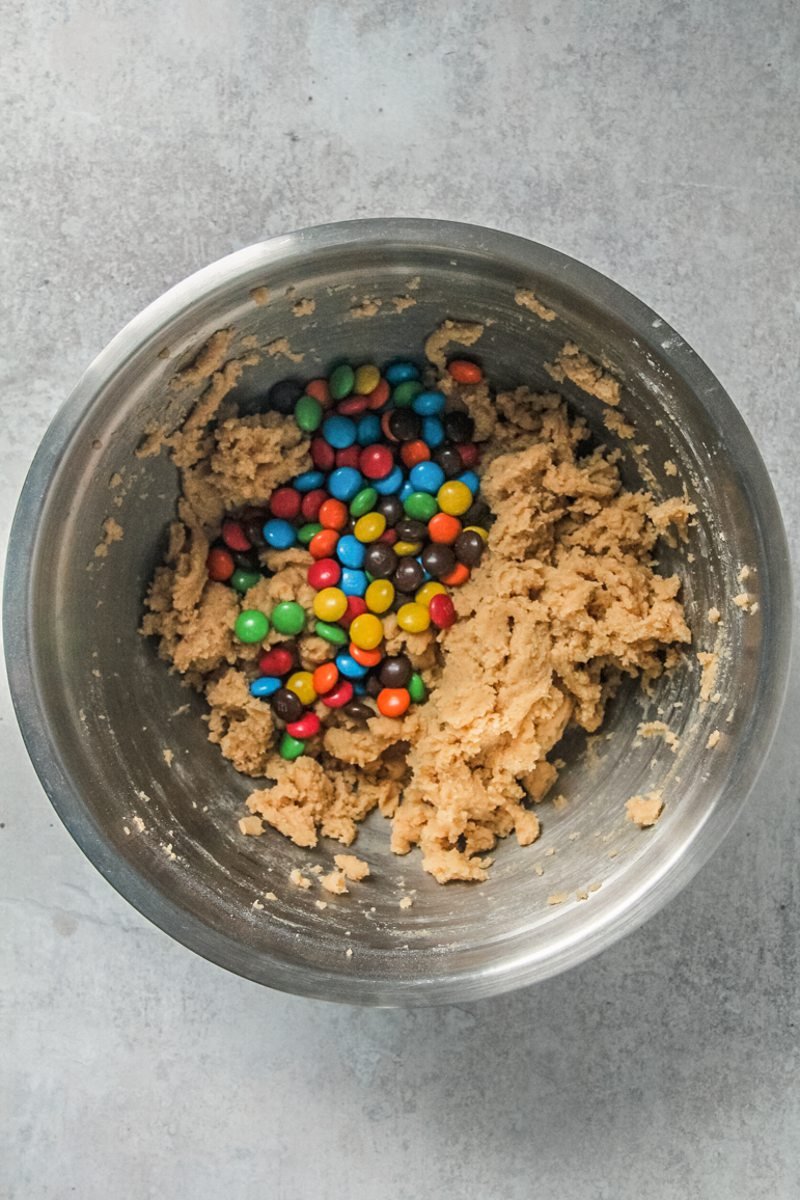 M&M's sit on top of cookie dough in a stainless steel bowl on a gray surface.