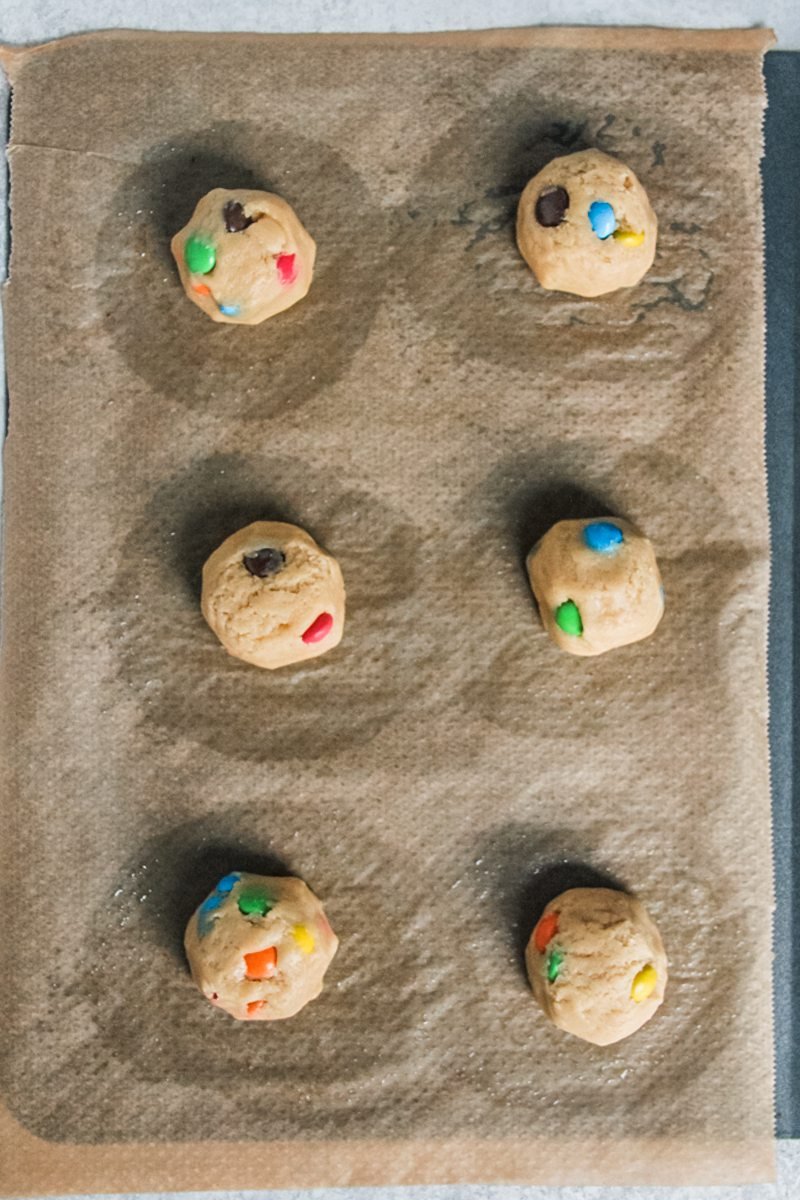 M&M cookie dough sits on parchment paper lined baking tray on a gray surface.