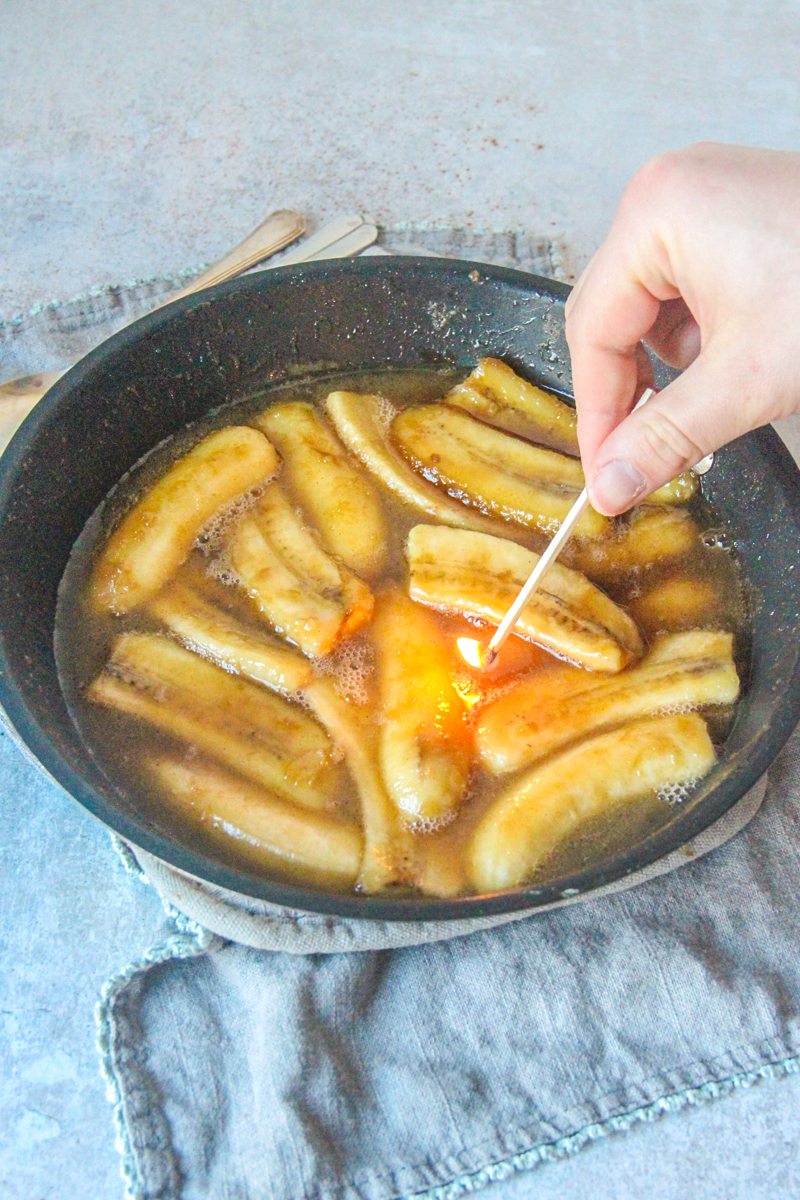 A match sits near Bananas Foster in a saucepan on a gray cloth on a gray surface.