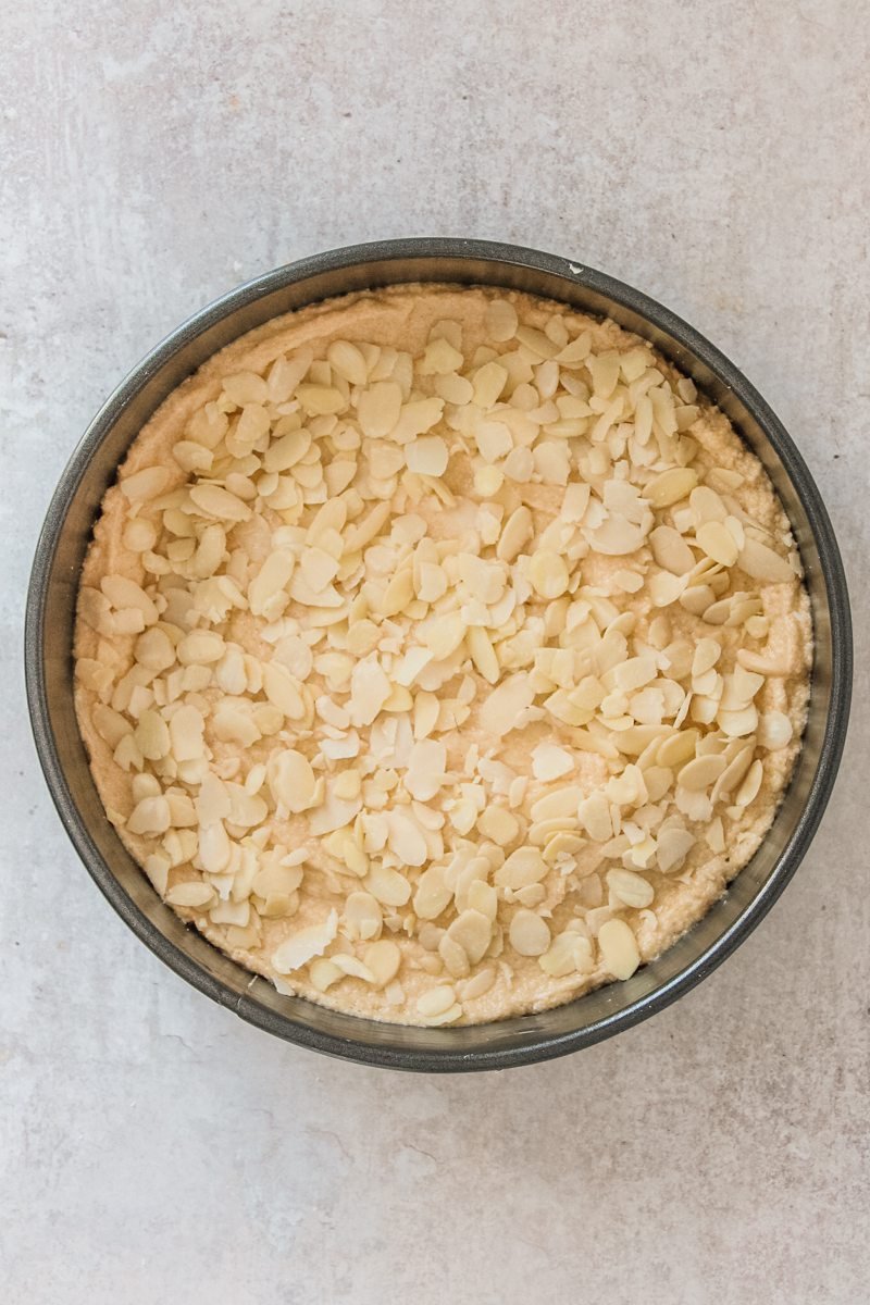 An unbaked Portuguese almond cake sits in a round metal cake tin on a gray surface topped with sliced almonds.