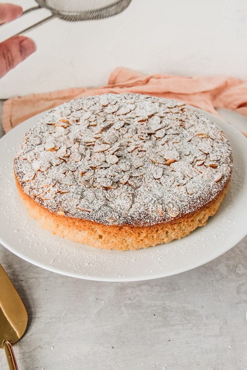 Powdered sugar is sprinkled over the top of a gluten free Portuguese almond cake on a gray surface.