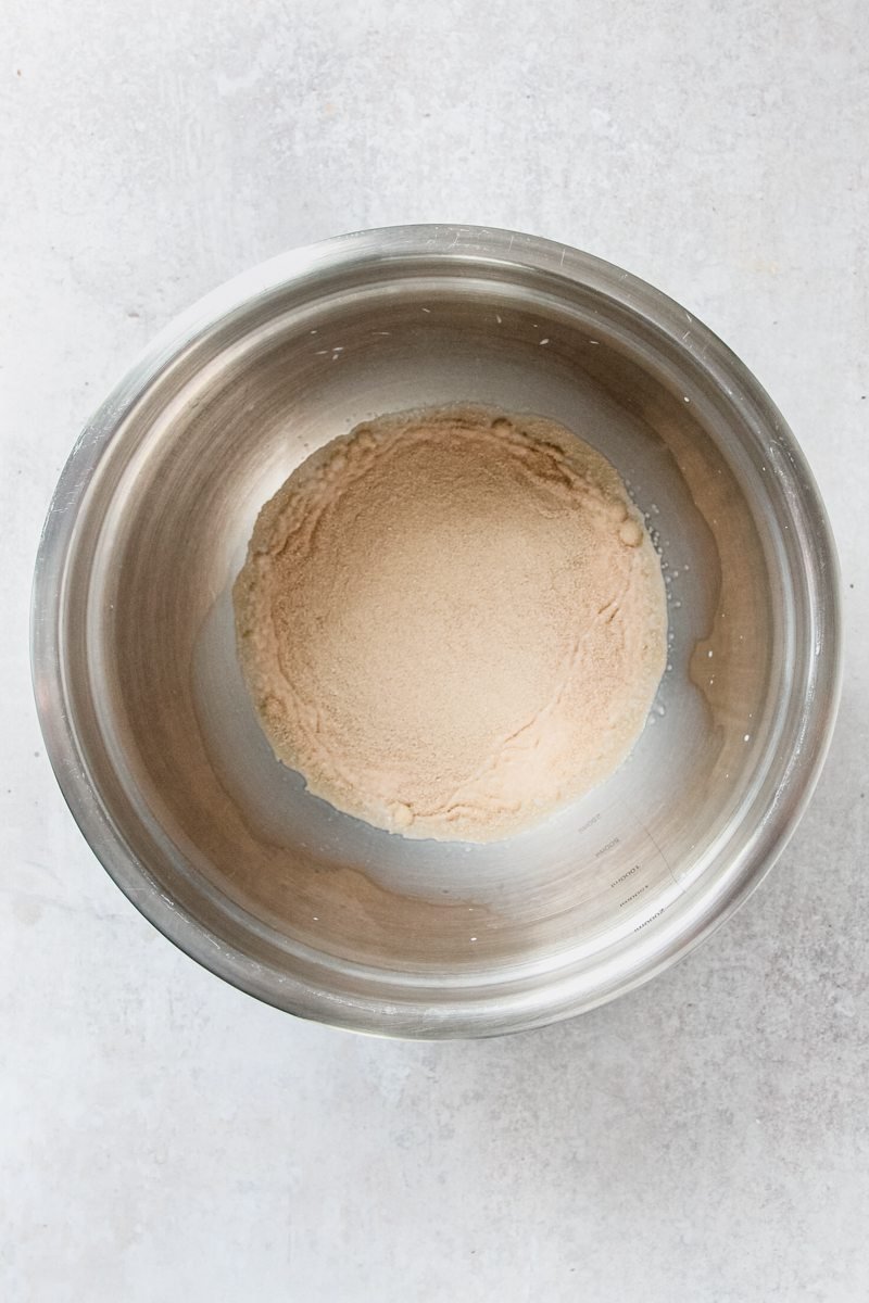 Yeast sits on top of warm milk in a stainless steel bowl on a gray surface.