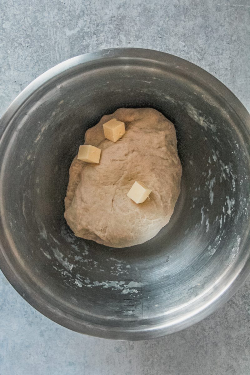 Cubes of butter sit on top of dough in a stainless steel bowl on a gray surface.