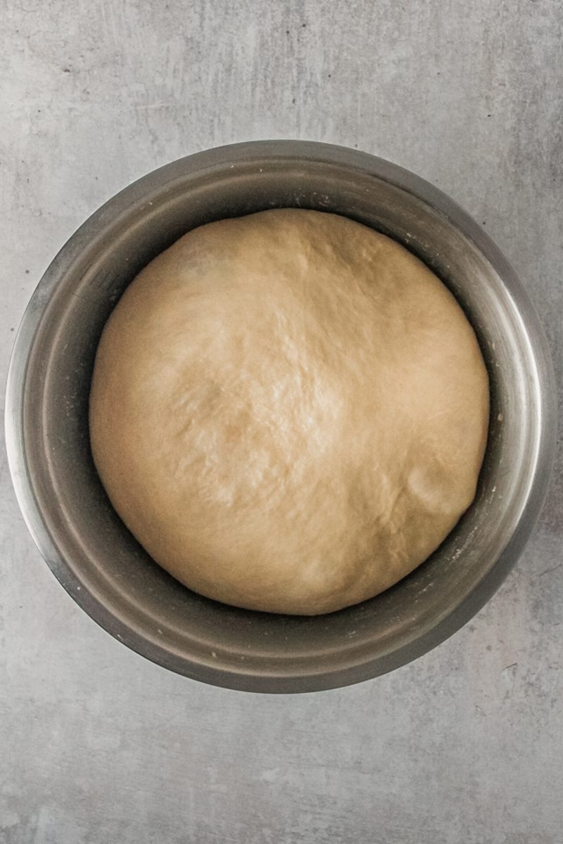 Dinner roll dough sits risen in a stainless steel bowl on a gray surface.