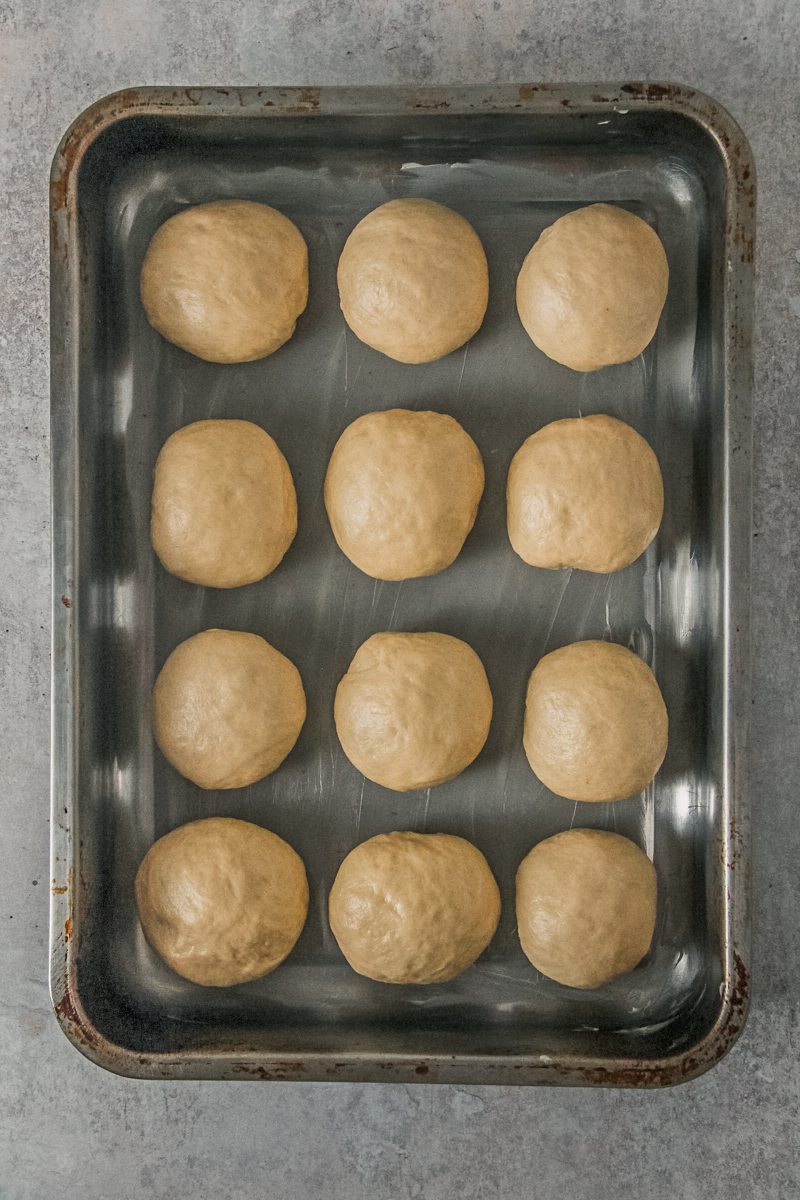 Unbaked dinner rolls sit in a rectangle metal baking tin on a gray surface for their second rise.