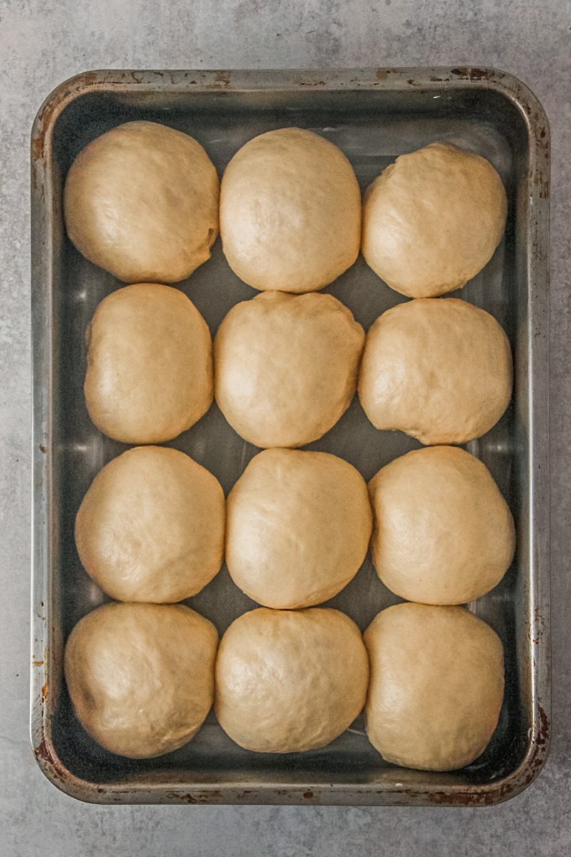 Fluffy unbaked dinner rolls sit in a rectangle metal baking tin on a gray surface.