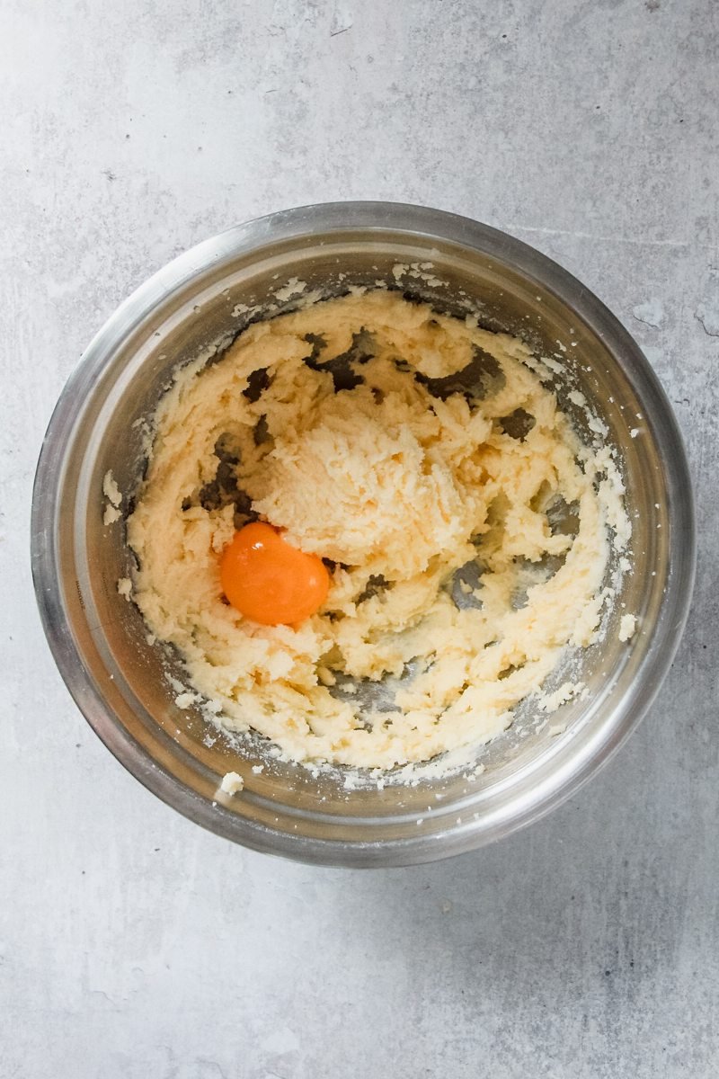 An egg yolk sits in a creamed butter and sugar mixture in a stainless steel bowl on a light grey surface.