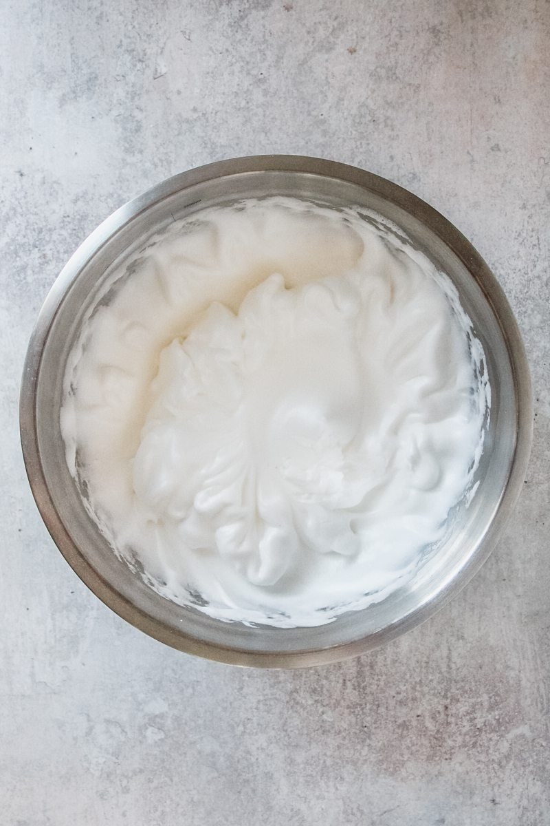 Whipped egg whites sit in a stainless steel bowl on a light grey surface.