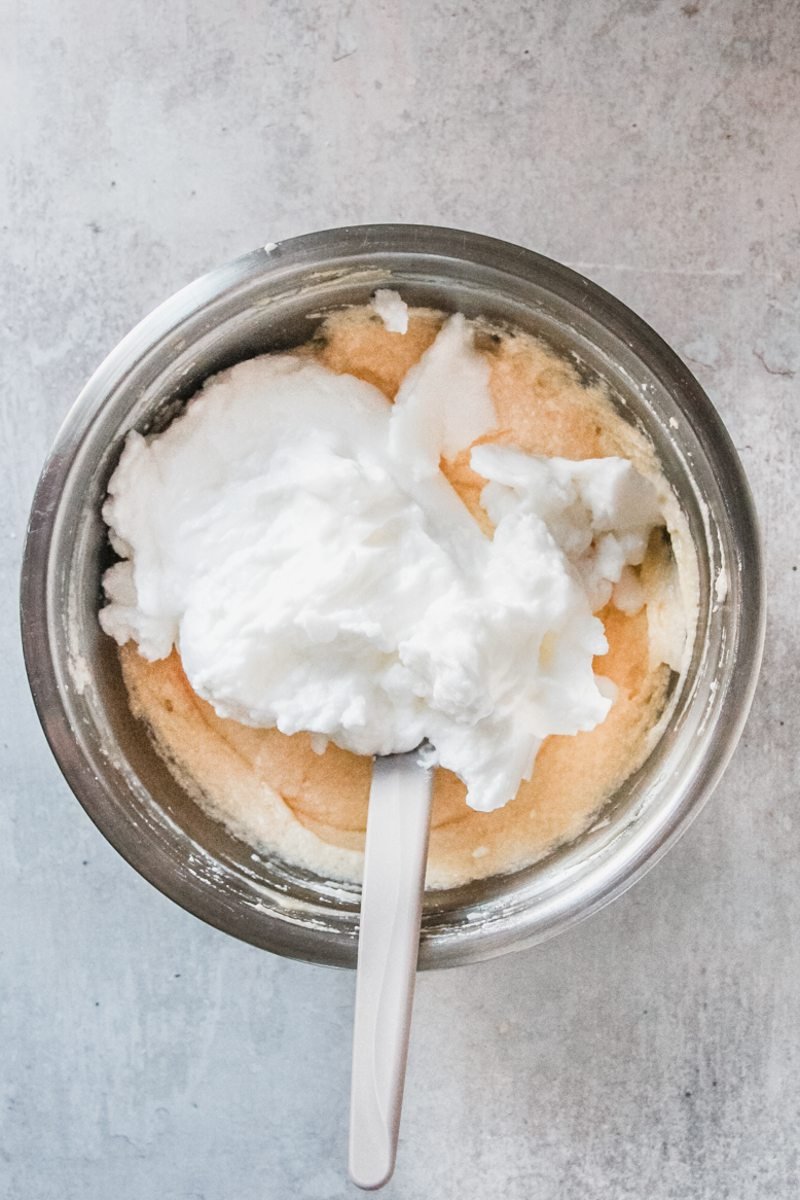 A portion of meringue mixture sits on top of a cake batter to be folded through for a Bublanina cake in a stainless steel bowl on a light grey surface.