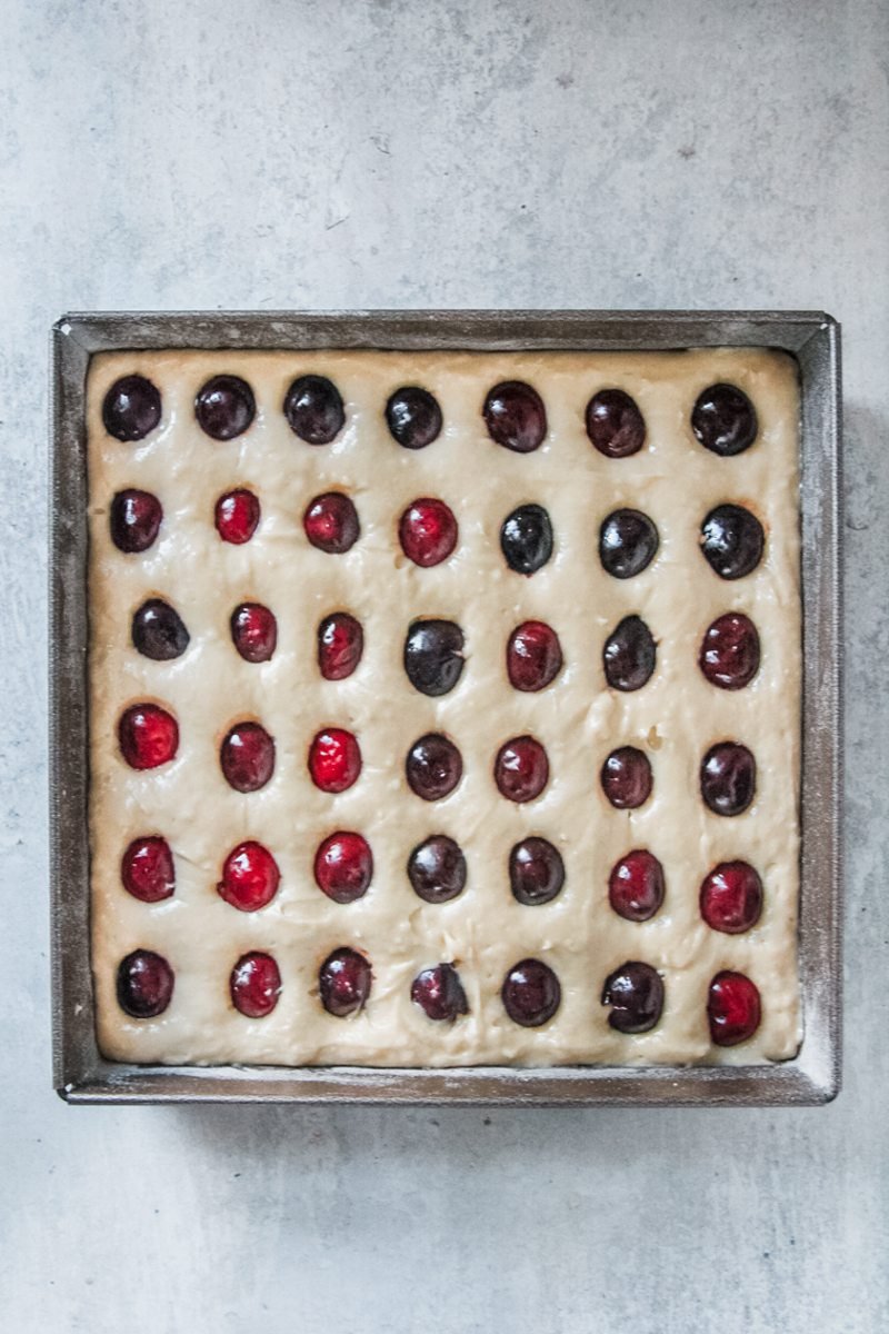 Pitted cherries speckle the cake batter in a square cake tin on a light grey surface.