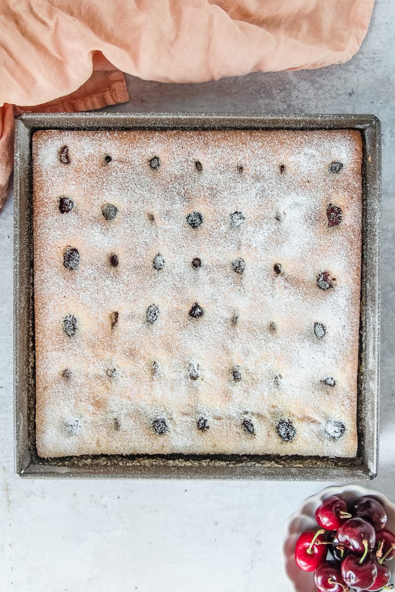 A Bublanina sits in a square tin with a little powdered sugar on top with a coral colored cloth beside the tin on a light grey surface.