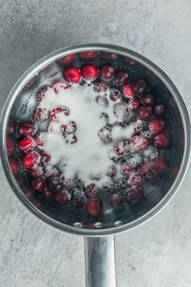 Cranberries and sugar sit in a stainless steel saucepan on a gray surface.