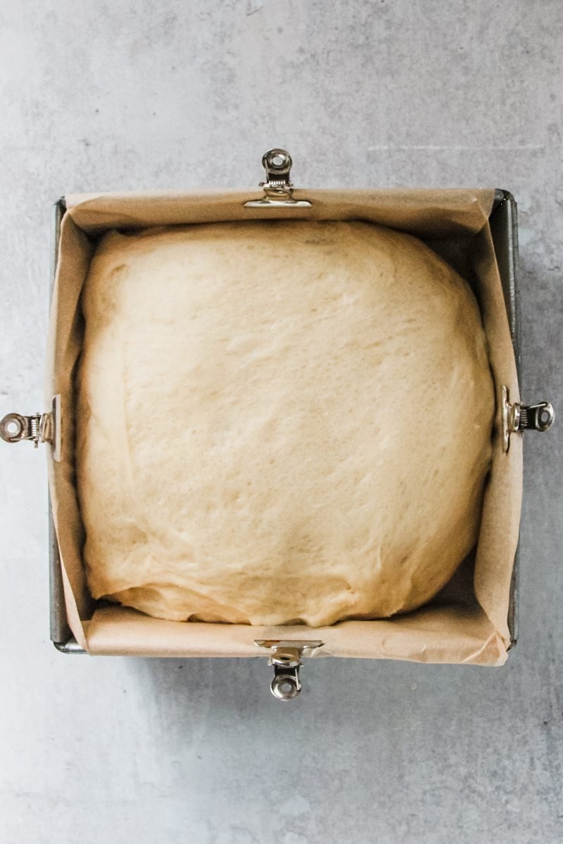 A yeasted cake brioche style dough sits in parchment lined square metal baking tin on a gray surface.