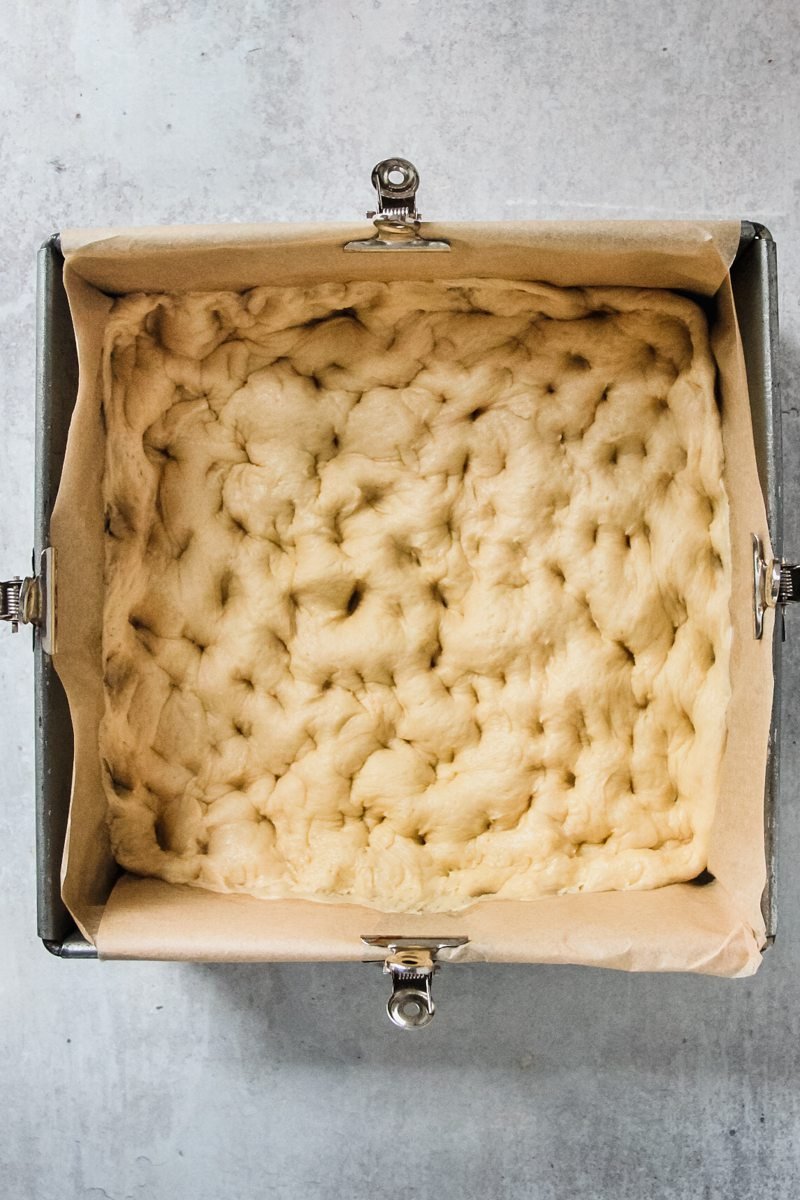 Brunsviger or Danish Coffee Cake yeasted dough with holes pocked all over it unevenly sits in a parchment paper lined metal baking tin on a gray surface.