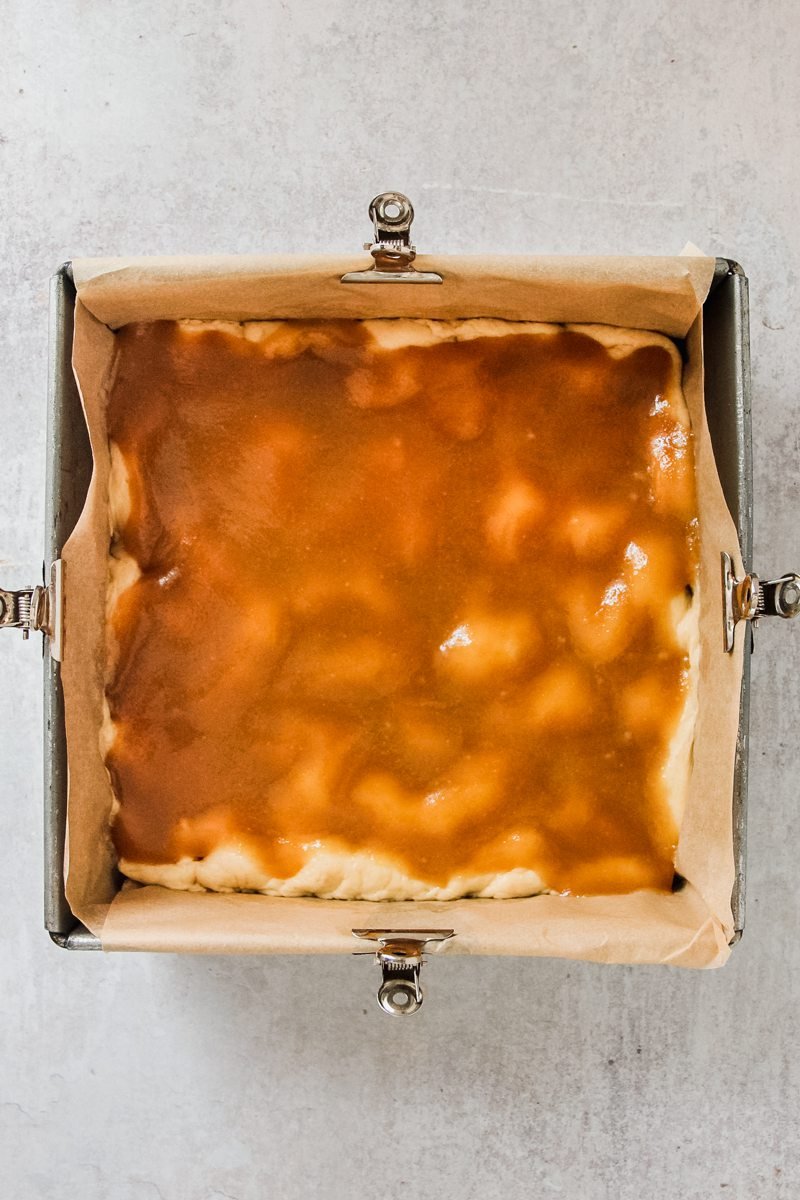 A Danish coffee cake dough sits in a parchment paper lined square metal tin with a caramel sauce on top of the uneven dough surface on a gray surface.