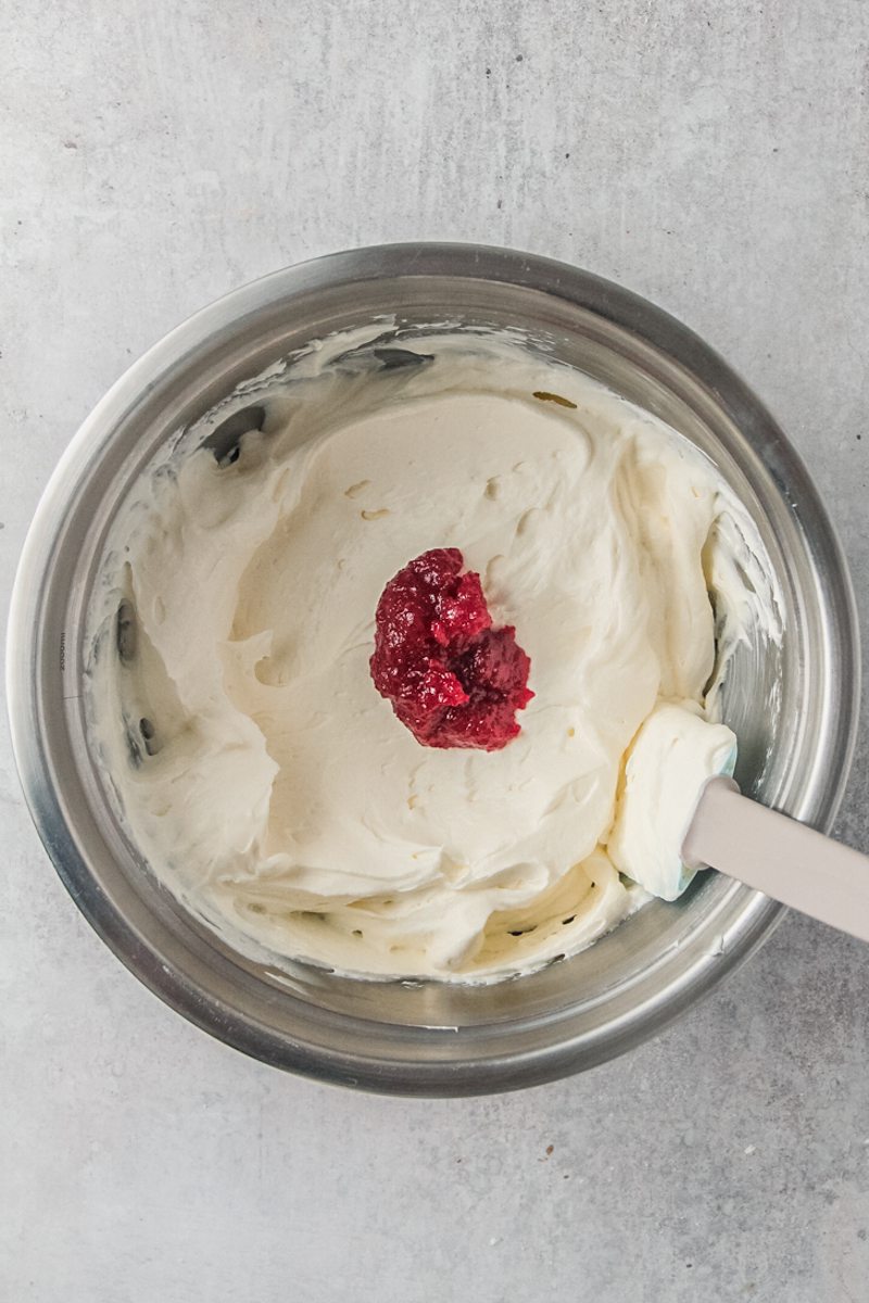 Cranberry puree sits in whipped cream cheese and cream in a stainless steel bowl on a gray surface.